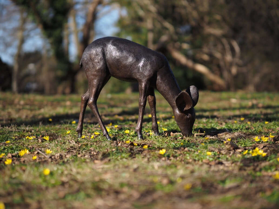 Bronze fawn sculpture grazing in a natural setting with flowers, showcasing detailed craftsmanship and outdoor durability.