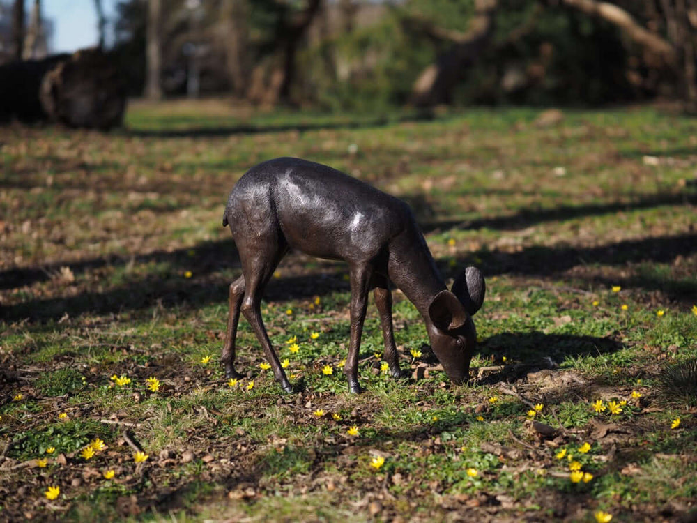 Bronze fawn sculpture grazing among flowers in a garden, showcasing detailed craftsmanship and outdoor durability.