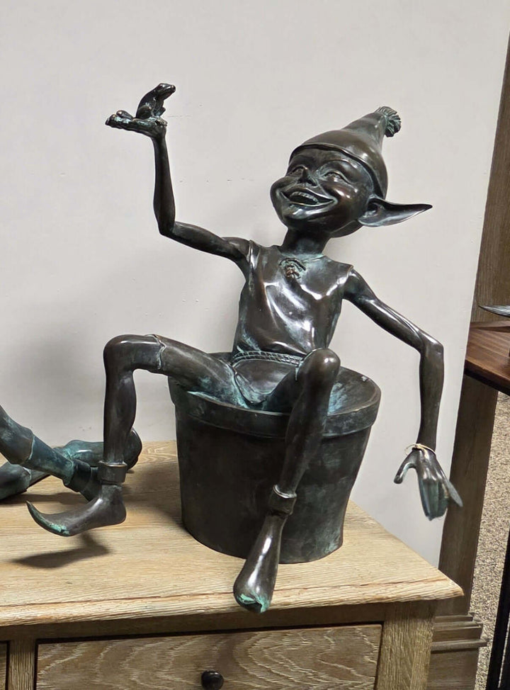 Bronze sculpture of a joyful pixie sitting in a bucket holding a frog, detailed craftsmanship with patina finish.