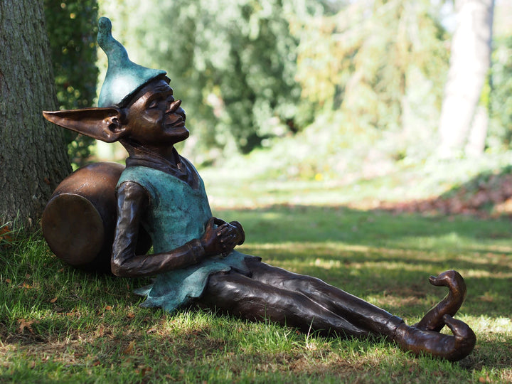 Joyful bronze pixie sculpture reclining against a barrel in a lush outdoor setting, showcasing intricate details and patina.