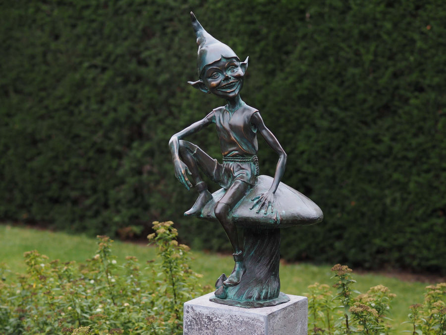 Bronze sculpture of a pixie with pointy hat sitting on a mushroom, finished with green patina, suitable for outdoors.
