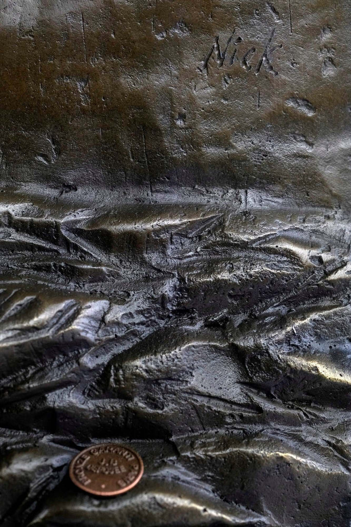 Close-up of bronze sculpture texture with artist signature 'Nick' and a coin for size comparison.
