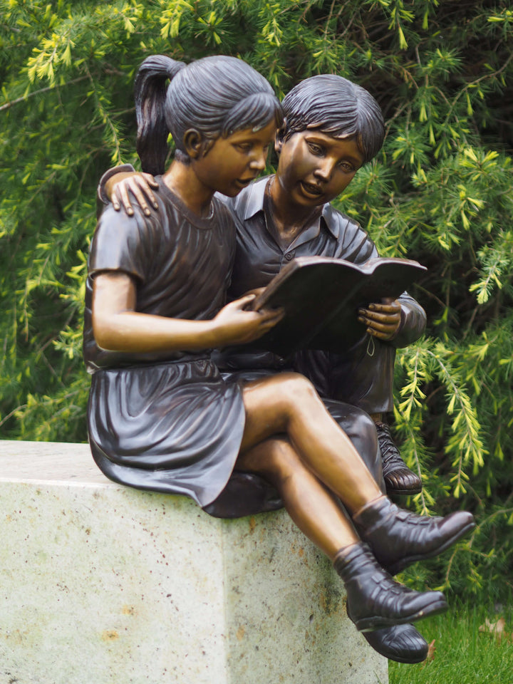 Bronze sculpture of a boy and girl reading together, showcasing friendship and childhood innocence in a garden setting.