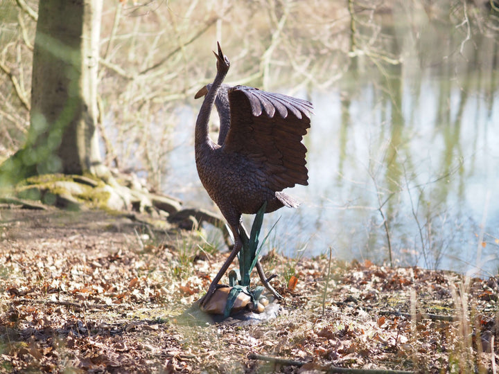 Elegant bronze stork fountain with wings spread, set by a tranquil water backdrop in a natural setting.