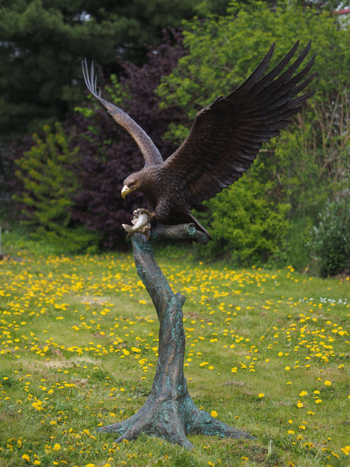 Large bronze eagle sculpture landing on tree trunk, wings spread wide, clutching fish in claws. Stunning detail.