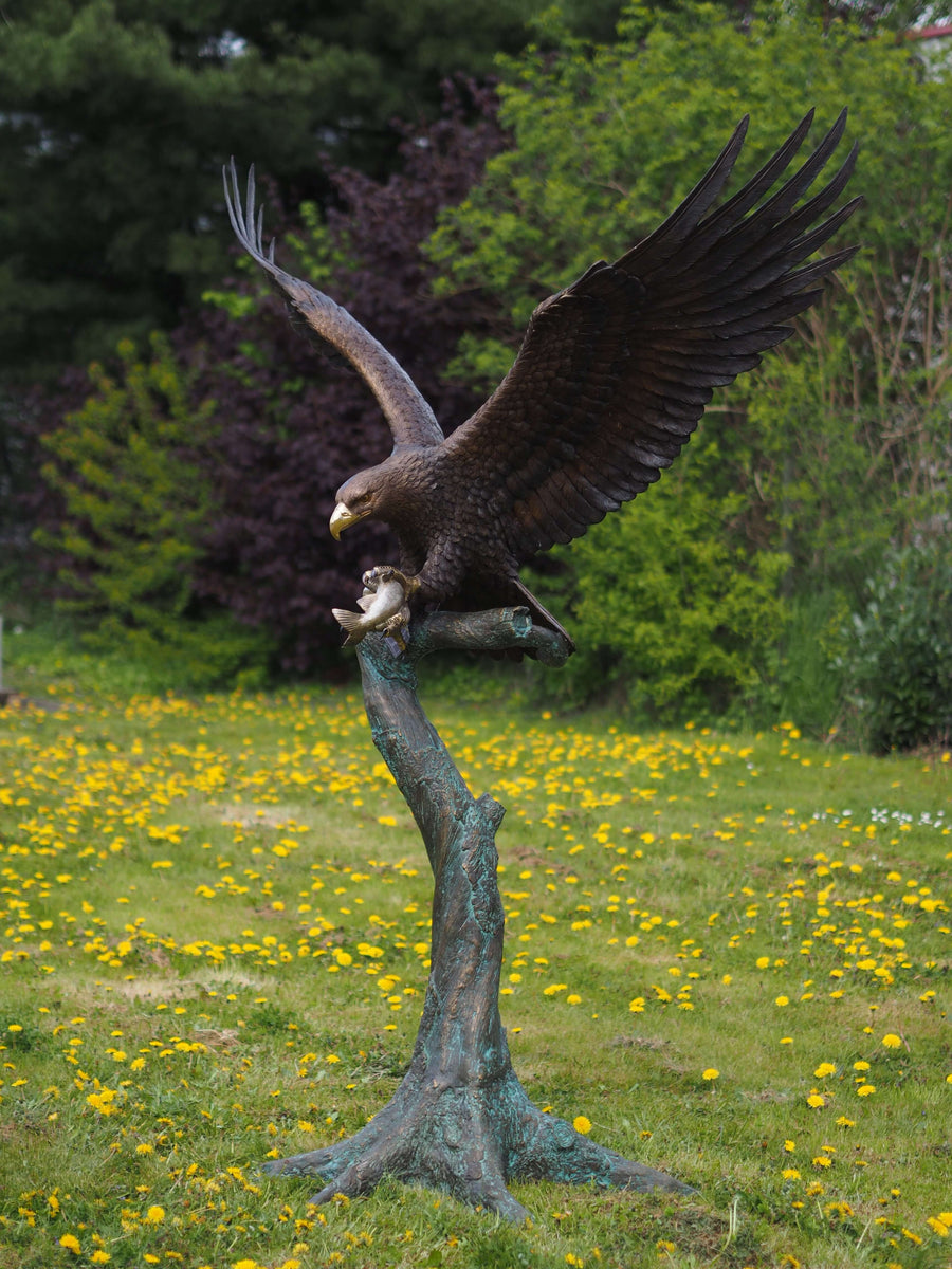 Large bronze eagle sculpture landing on tree trunk, wings spread wide, clutching fish in claws. Stunning detail.