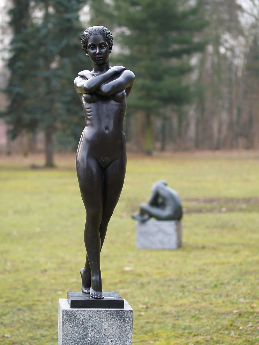 Large bronze sculpture of a standing nude woman with arms crossed, showcasing elegance in a serene outdoor setting.