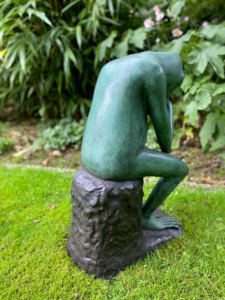 Bronze sculpture of a thinking figure sitting on a stone, surrounded by greenery in a garden.